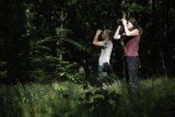 Zwei Personen stehen auf einer Waldlichtung und schauen mit Ferngläsern nach oben.