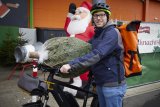 Ein Mann steht mit einem E-Bike vor einer Verkaufsstelle für Weihnachtsbäume. Einen verpackten Weihnachtsbaum hat er auf Lenker und Sattel abgelegt.